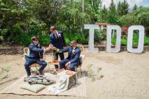 A group of groomsmen sitting on the sand with a bottle of wine.