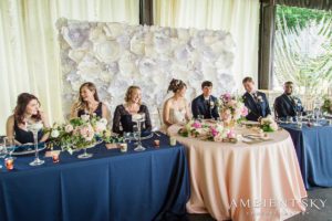 A wedding party sits at a table in front of a blue tablecloth.