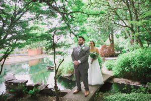 A couple, dressed as bride and groom, standing on a picturesque bridge near a serene pond.