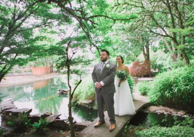 A couple, dressed as bride and groom, standing on a picturesque bridge near a serene pond.