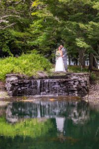 A bride and groom standing next to a pond in a shady grove.